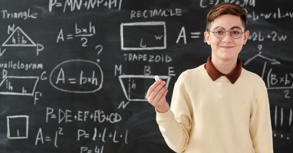 Young boy holding chalk in front of blackboard with math formulas, wearing glasses and smiling.